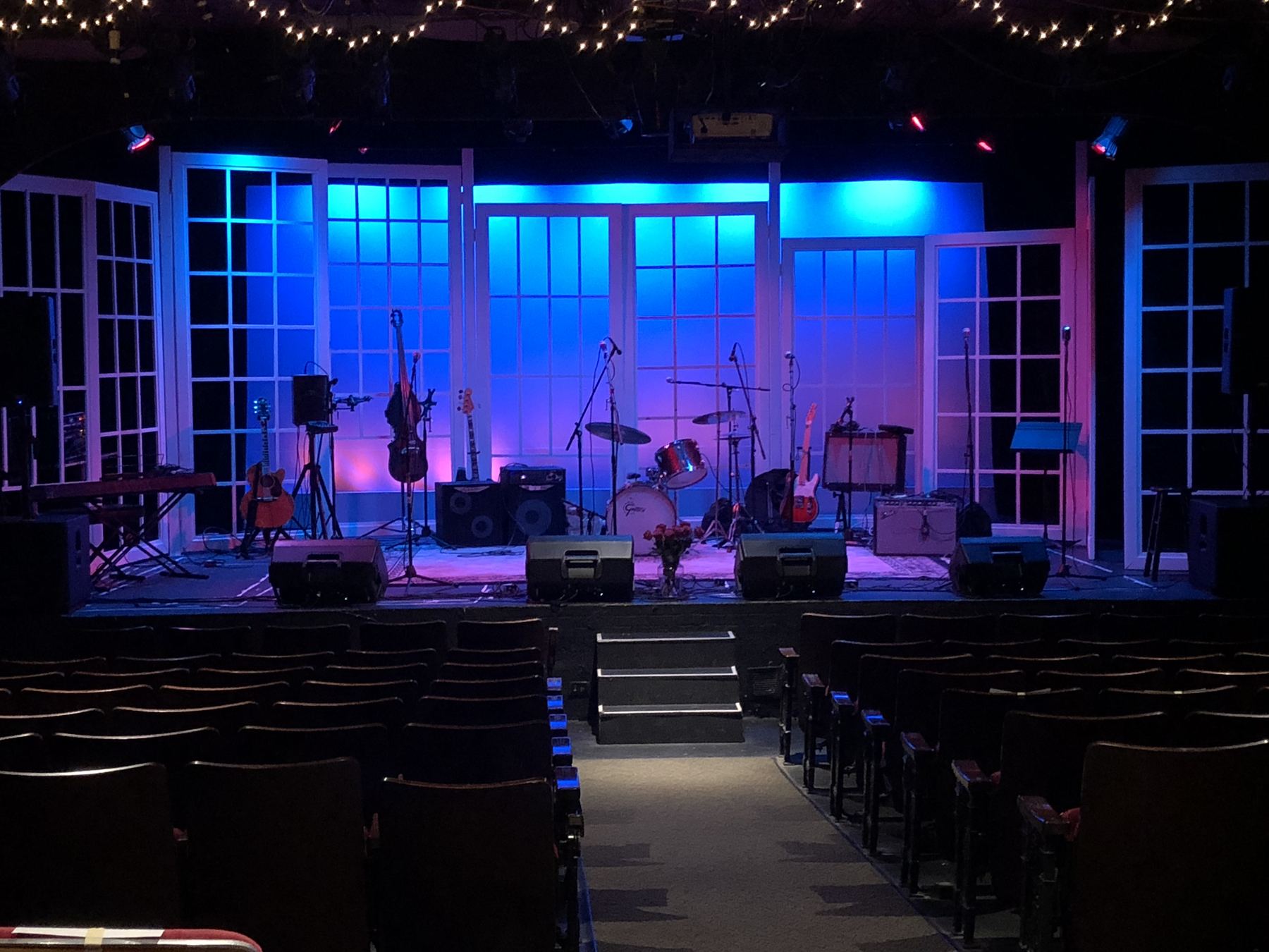 A stage set up for a band, lit in blue and pink, with monitors and instruments ready